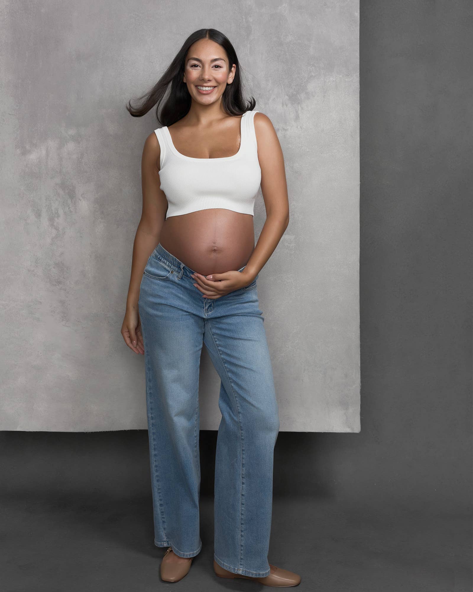 Pregnant woman wearing a white top and blue jeans against a gray background