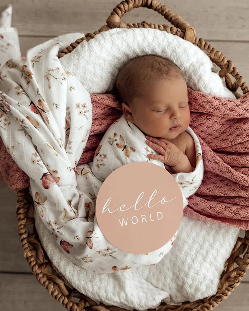 Newborn baby wrapped in a floral blanket with a 'hello world' card in a wicker basket.
