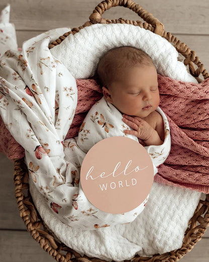 Newborn baby wrapped in a floral blanket with a 'hello world' card in a wicker basket.